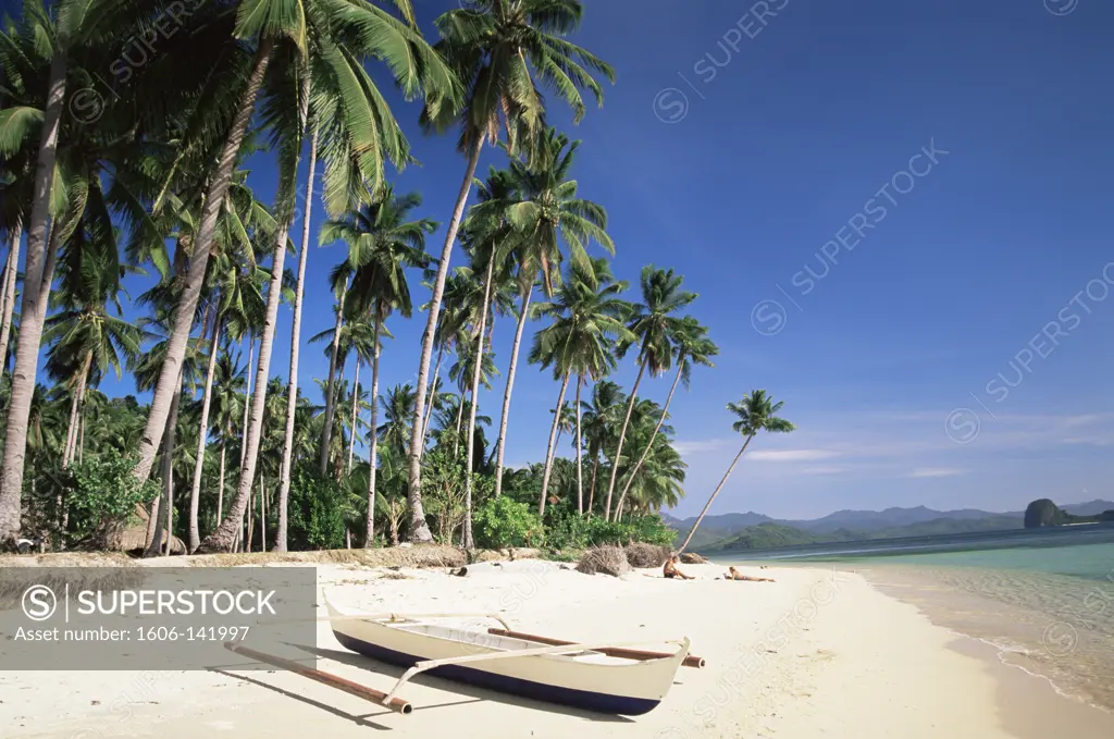 Philippines,Palawan,Bascuit Bay,El Nido,Couple Sunbathing on Beach with Outrigger Boat in Foreground