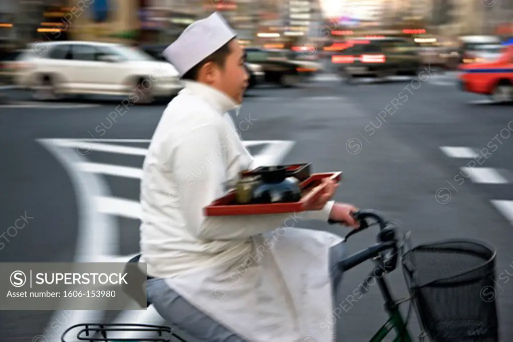A young Japanese restaurant worker on a bicycle crosses Aoyama Avenue while delivering an order in the trendy yet upscale Omotesando District of Tokyo, Japan.