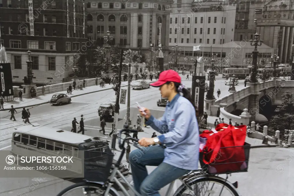 A young Japanese woman uses her cellphone while riding a bicycle past an old 1950's black-and-white mural of Nihonbashi Bridge at a construction site in the Nihonbashi District of central Tokyo, Japan.
