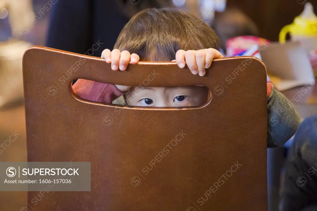 A shyly curious young Japanese boy peers through an opening in the back of his chair at a cafe inside the new Kyoto Station Building in Kyoto, Japan.
