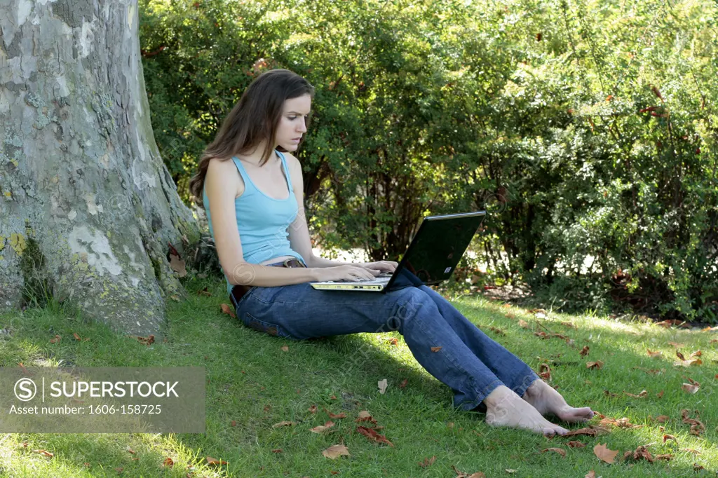France,Ile de France. Young woman sitting in the park under a tree and using her laptop