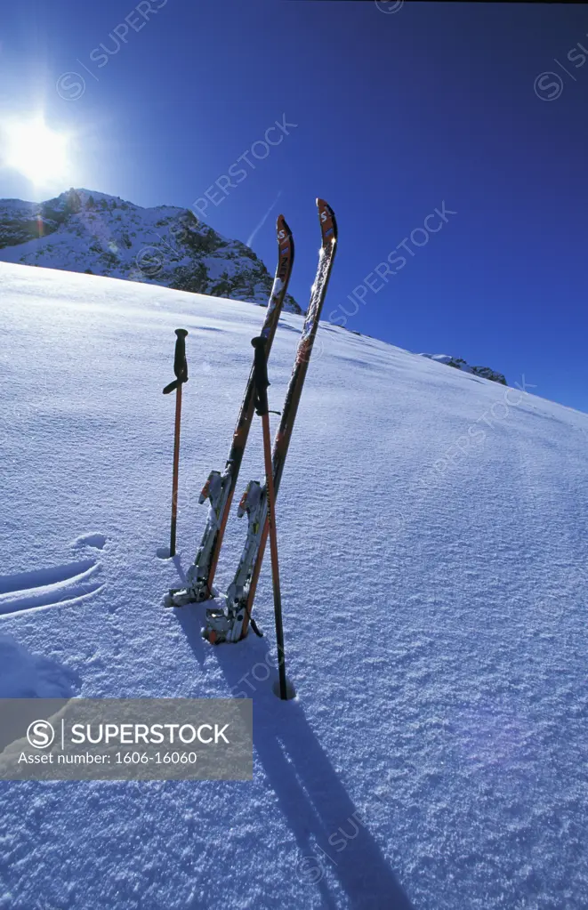 Pair of skis and poles planted in powder snow, mountains in the background, blue sky