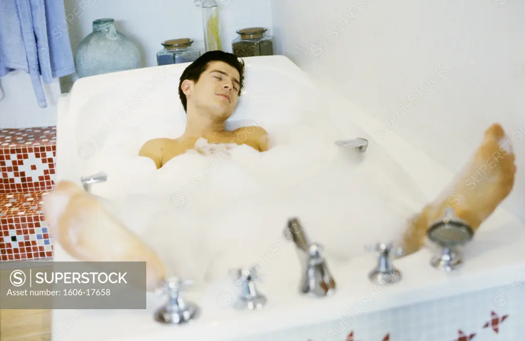 Young man lying in a bathtub full of foam, feet sprayed outside of water