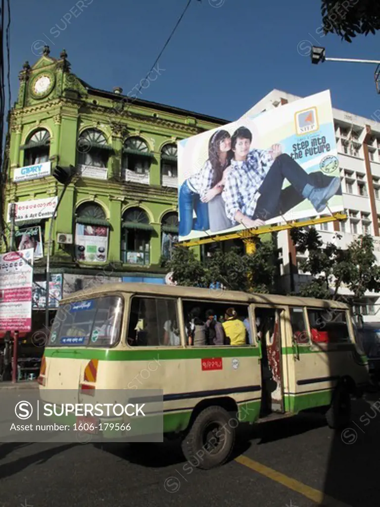Myanmar Burma Yangon an old bus is passing under a billboard showing 2 teenagers in front of an old colonial green  building