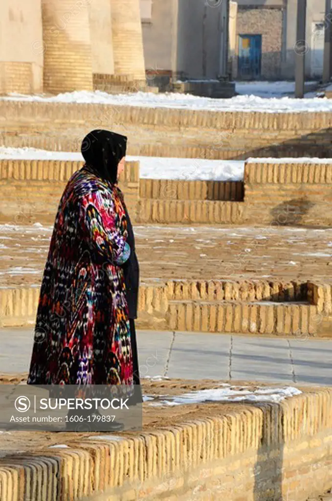 /UZBEKISTAN KHIVA a woman in a long colorful coat is standing in front of the KUNYA ARK fortress