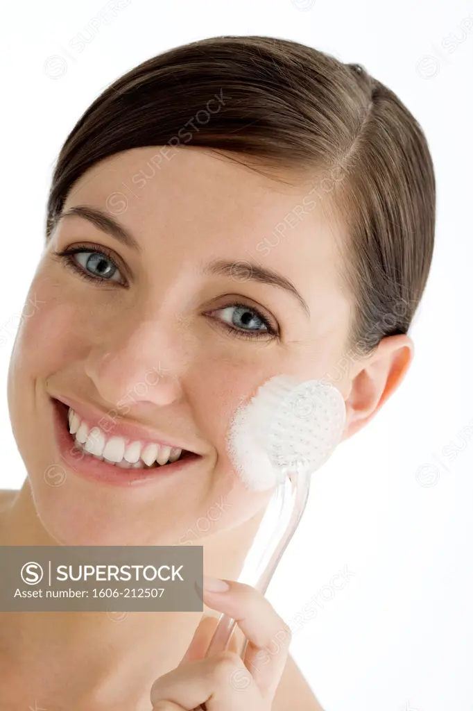 Portrait of a smiling young woman using a brush to remove her make-up on her cheek, looking at camera