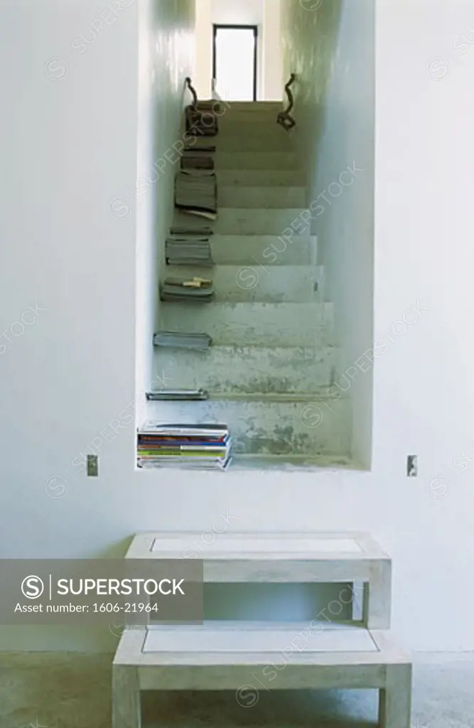 Intérieur maison blanche, 2 marches en bois devant escalier, piles de magazines sur marches, porte vitrée en haut