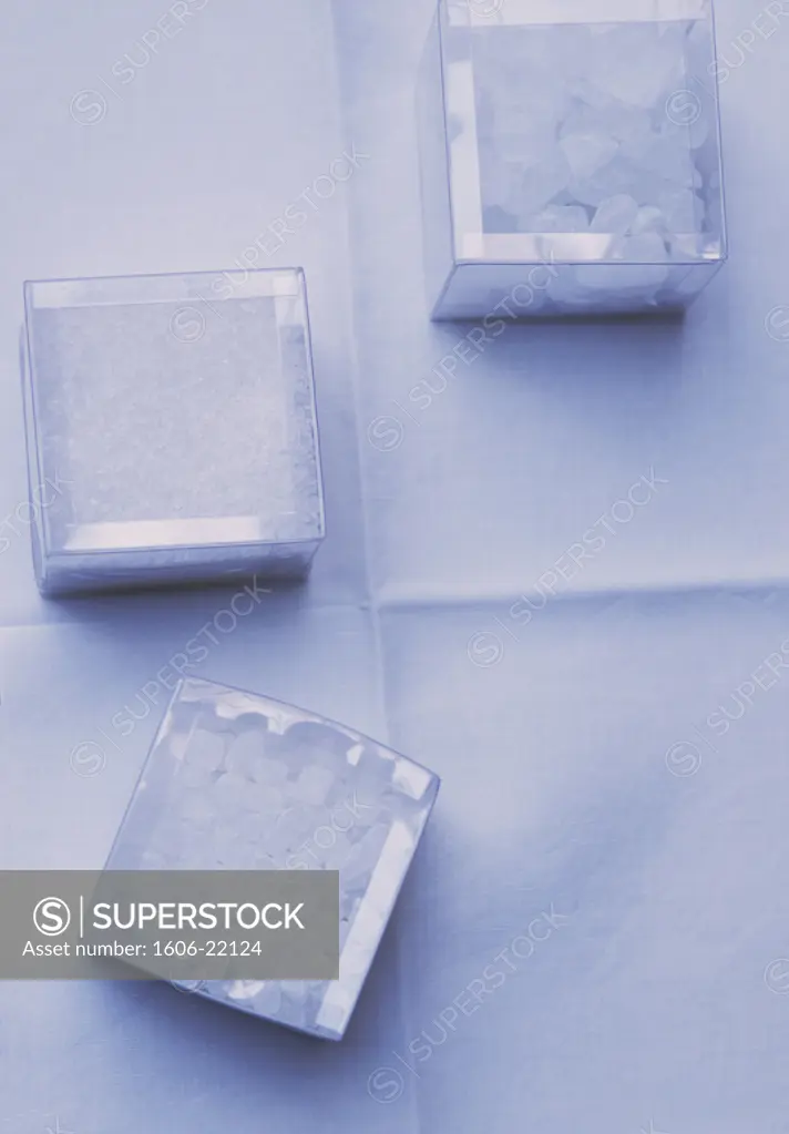 Vue plongeante sur cristeaux de sucre candi dans 3 petites boites carrées transparentes, sur tissu blanc