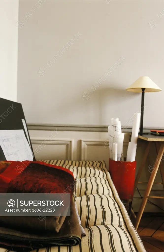 Intérieur bureau d'architecte, couverture et tissus pliés sur banquette en tissu rayé, plans roulés dans boîte rouge, lampe, murs blancs