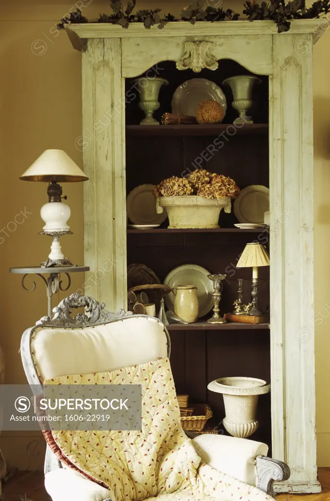 Intérieur salon ancien, bibelots sur étagères d'un meuble en bois, fauteuil au 1er plan, lampe sur sellette