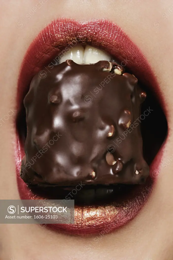 IN*Gros plan sur bouche d'une jeune femme tenant rocher au chocolat entre ses dents, lèvres maquillées