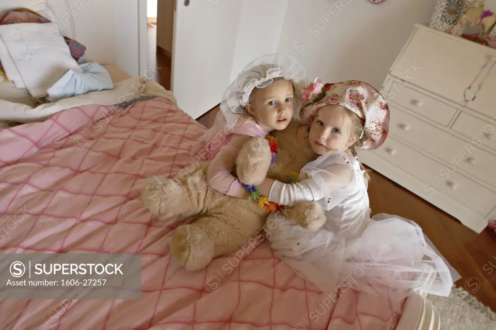 Plunging view on two girls posing, with hats and princess dresses, hugging teddy bear, on a bed