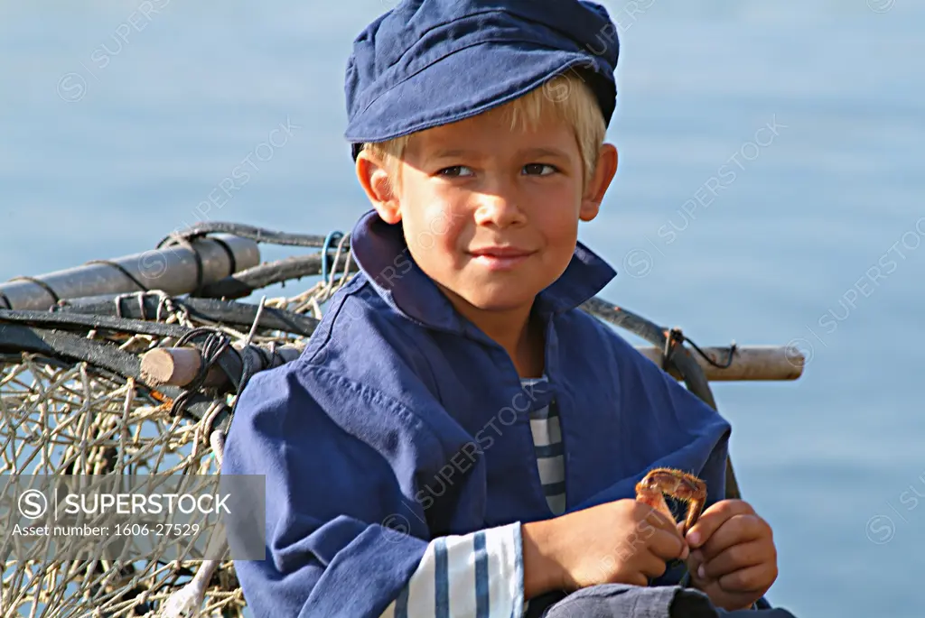 Boy smiling, sitting on dockside, conical fishing basket, jacket and and sailor hat