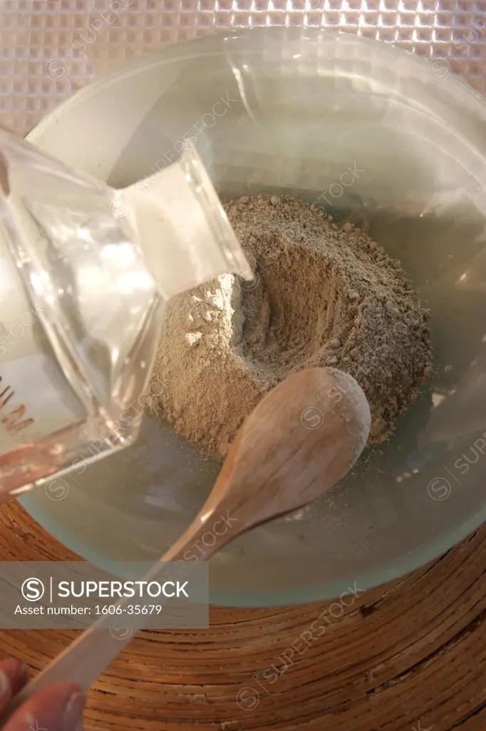 Preparation of an exfoliating face-scrub with clay, in glass bowl, water running from. a bottle, spatula, white towel