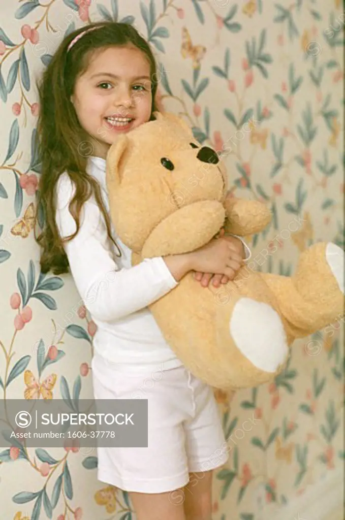 Little girl posing, standing in a room, hugging big teddy bear, wallpaper