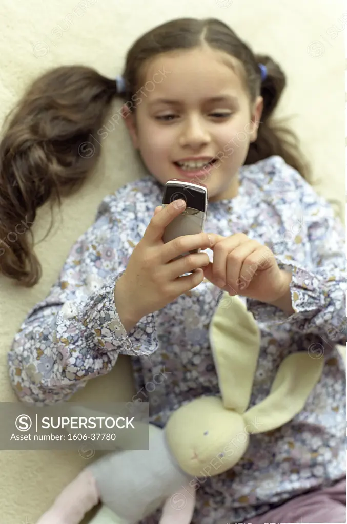 Plunging view on little girl smiling, looking at the screen of her mobile, plush rabbit, carpet