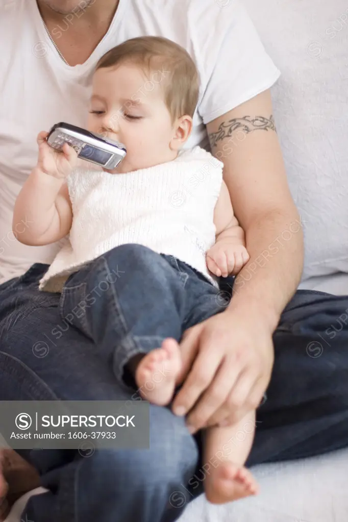 Little boy sitting on man's knees on a sofa, white sleeveless sweater, jeans, playing with mobile phone