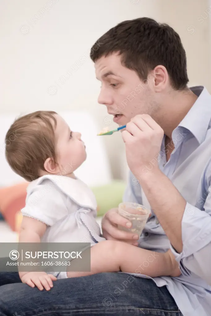 Man feeding baby seated on his knees, profile, glass pot, spoon