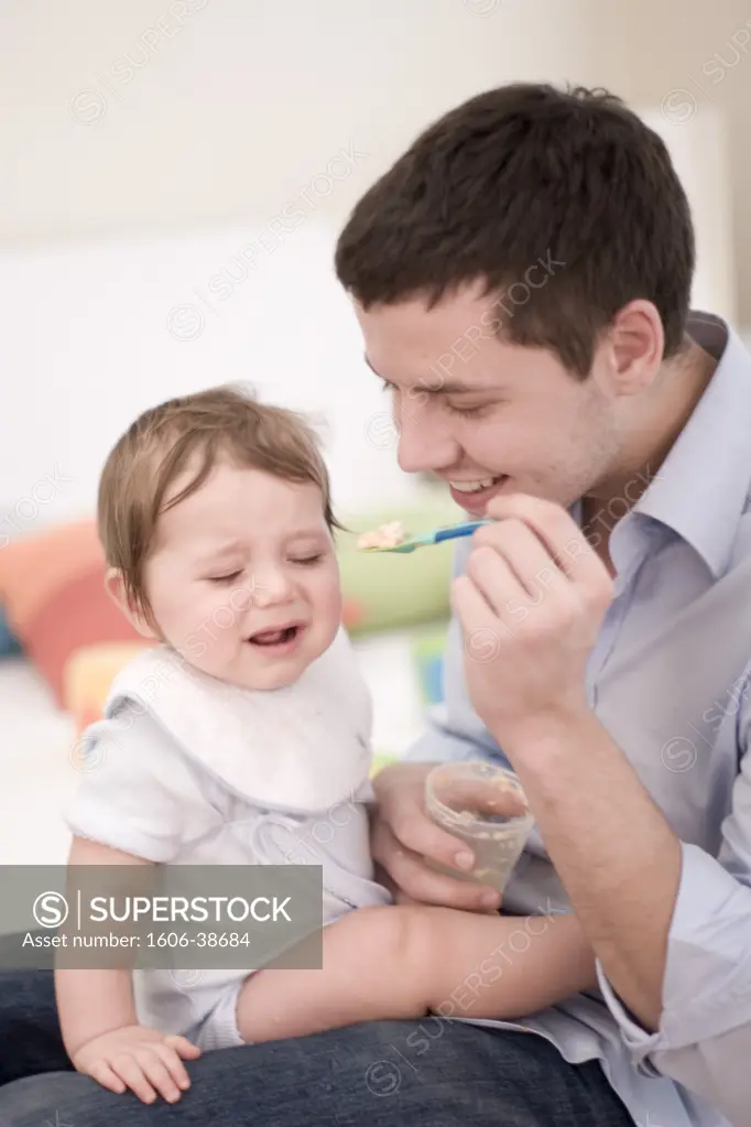 Man feeding baby seated on his knees, crying, glass pot