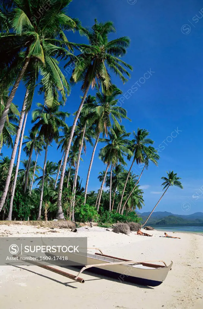 Philippines, Palawan, Bascuit Bay, El Nido, Couple Sunbathing on Beach with Outrigger Boat in Foreground