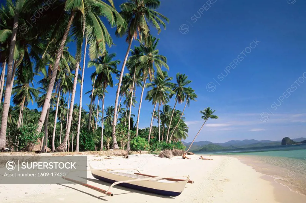 Philippines, Palawan, Bascuit Bay, El Nido, Couple Sunbathing on Beach with Outrigger Boat in Foreground