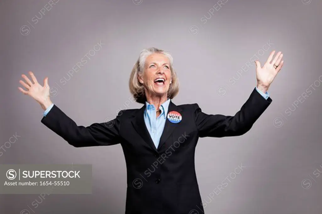 Studio. Senior businesswoman wearing Vote Badge with arms raised on grey background