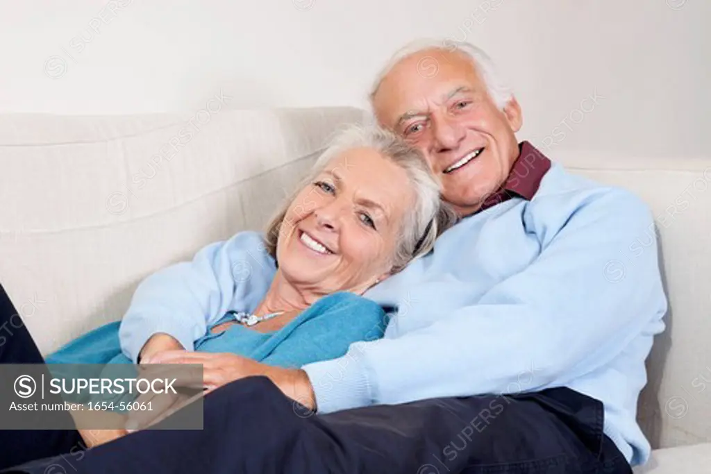 London, UK. Portrait of happy elderly man with arm around spouse lying on sofa at home
