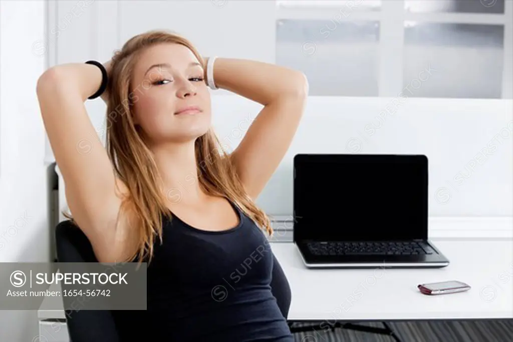 London, UK. Portrait of young businesswoman with hands behind head sitting in front of desk in office