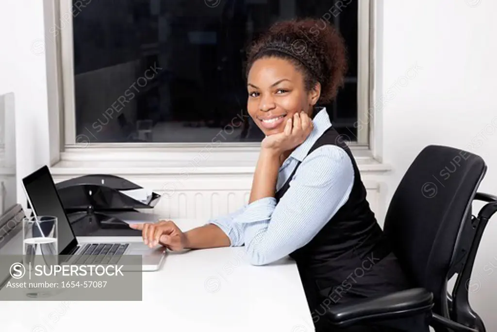 London, UK. Portrait of smiling young businesswoman using laptop at desk in office