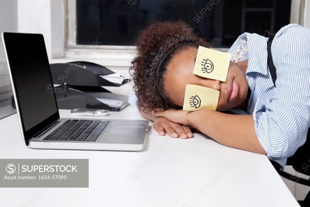 London, UK. Businesswoman sleeping with sticky notes on eyes at desk in office