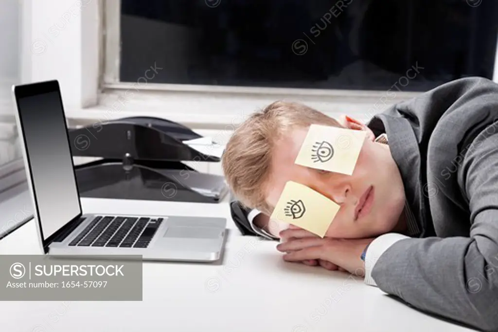 London, UK. Businessman sleeping with sticky notes on eyes at desk in office