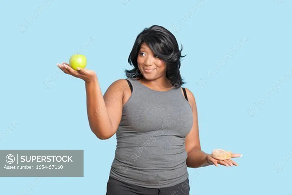 London, UK. Overweight mixed race woman holding green apple and donut over blue background