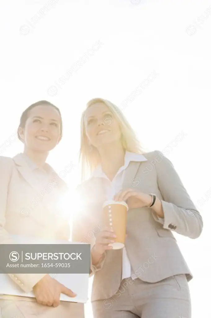 Happy young businesswomen with disposable cup and laptop against sky