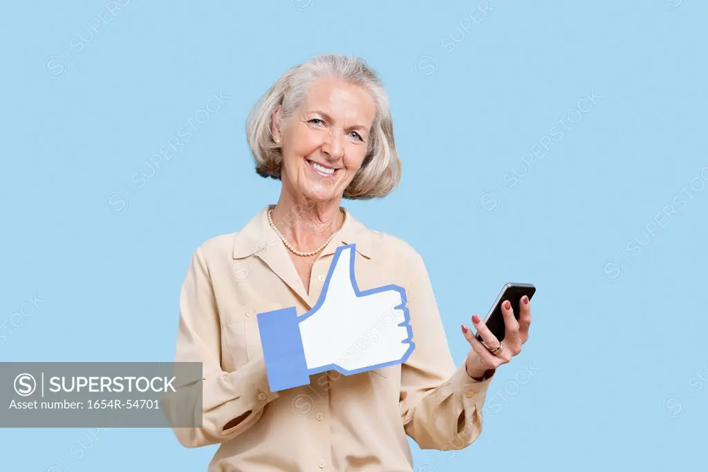 Portrait of senior woman with cell phone holding fake like button against blue background