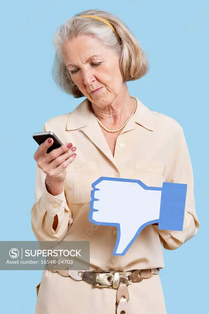Senior woman with cell phone holding fake dislike button against blue background