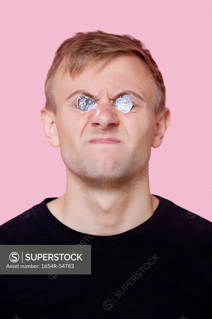 Funny young man covering eyes with coins over pink background