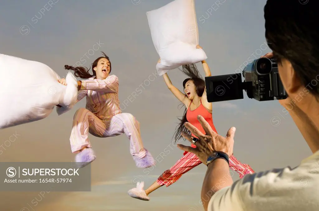 Man video recording two playful women in sleepwear having a pillow fight