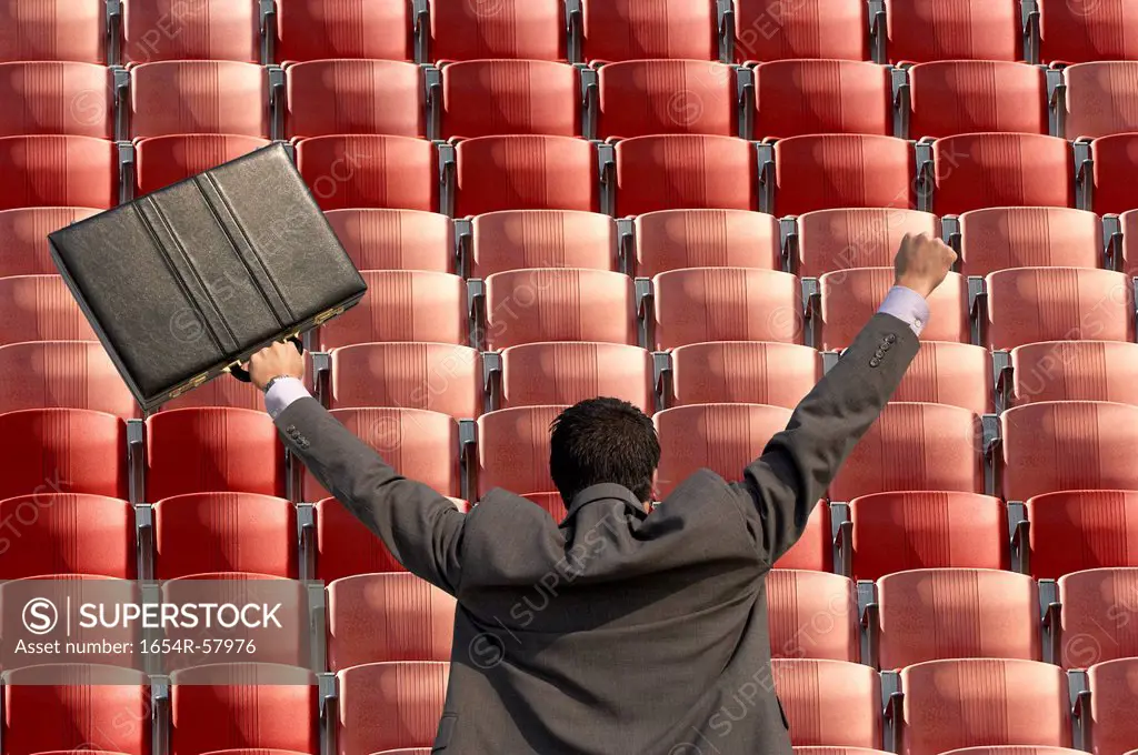 Back view of victorious businessman with briefcase facing rows of red seats at stadium