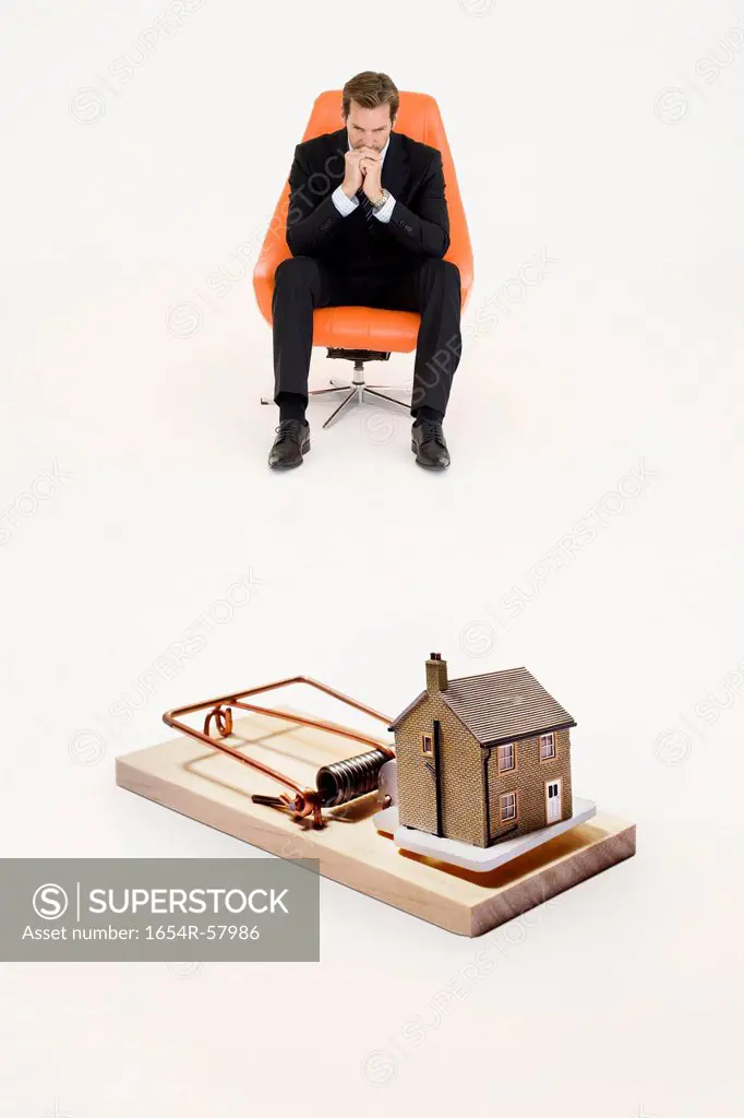 Model home on mouse trap with worried businessman sitting on chair representing increasing real estate rates