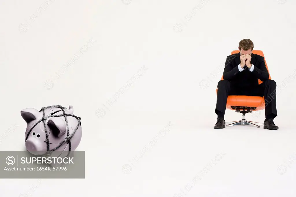 Worried businessman on chair and piggybank tied with rope representing financial difficulties