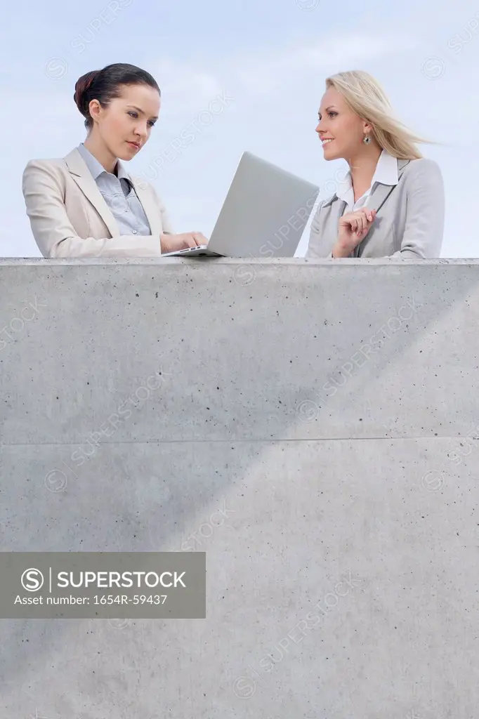 Low angle view of businesswoman using laptop while standing with coworker on terrace against sky