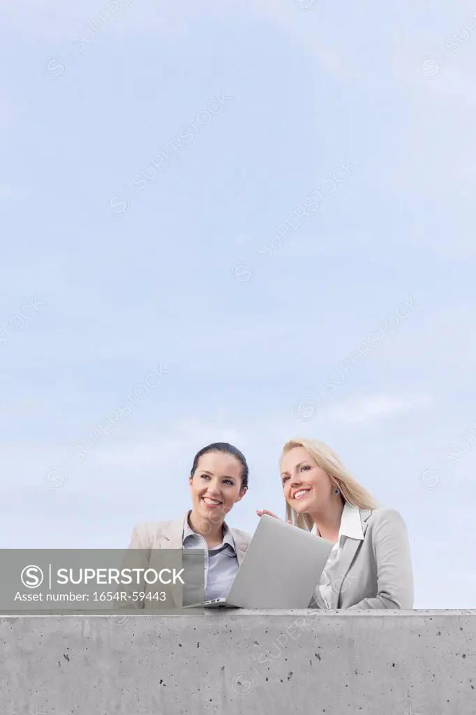 Low angle view of happy young businesswomen with laptop looking away while standing on terrace against sky