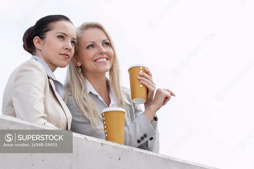 Low angle view of businesswomen with disposable coffee cups looking away while standing on terrace against sky