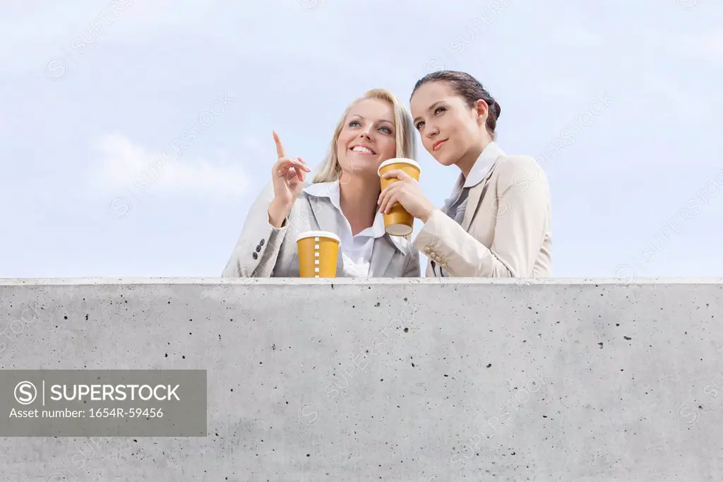 Low angle view of young businesswomen with disposable coffee cups looking away while standing on terrace against sky