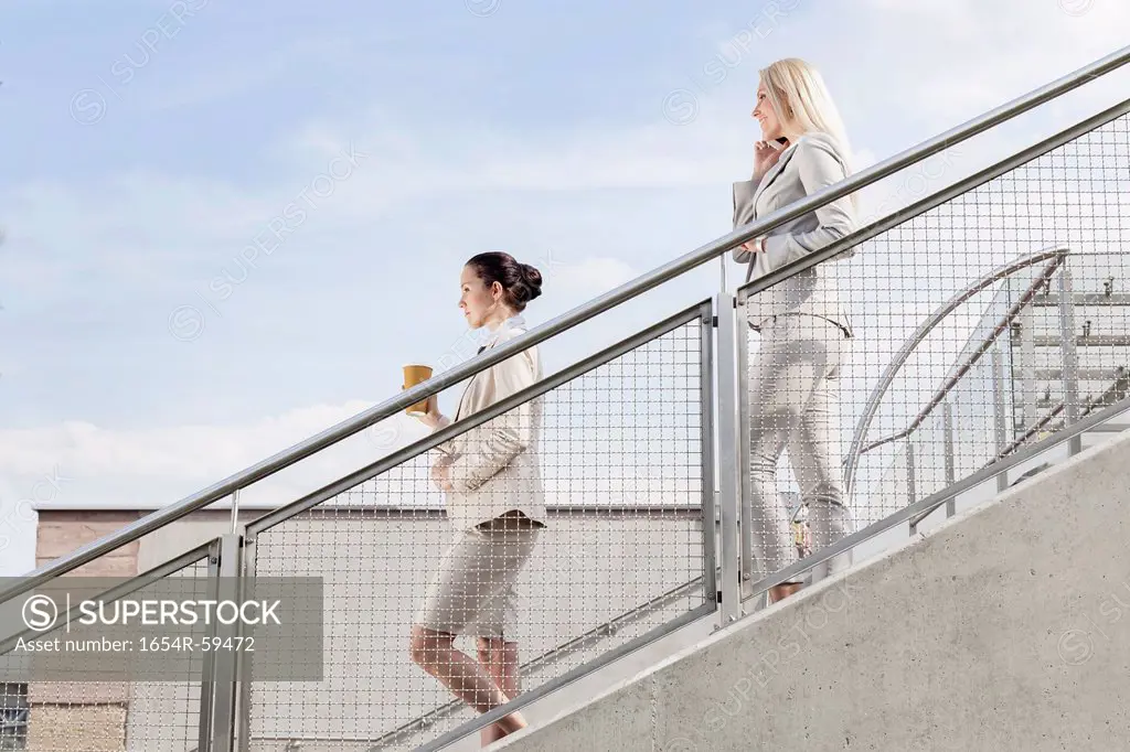 Profile shot of businesswomen holding disposable cup and mobile phone while moving down stairs together against sky