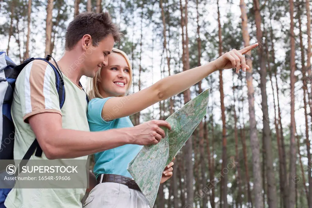 Happy young hiking couple holding map with woman pointing away in forest