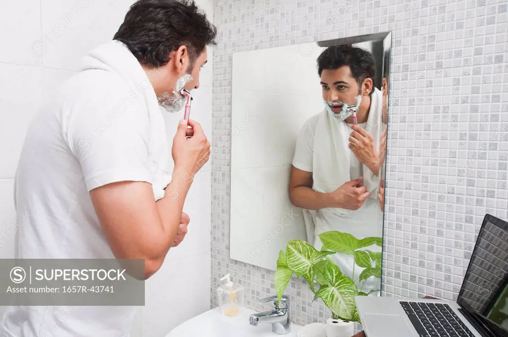 Man shaving his face in the bathroom with a laptop beside him