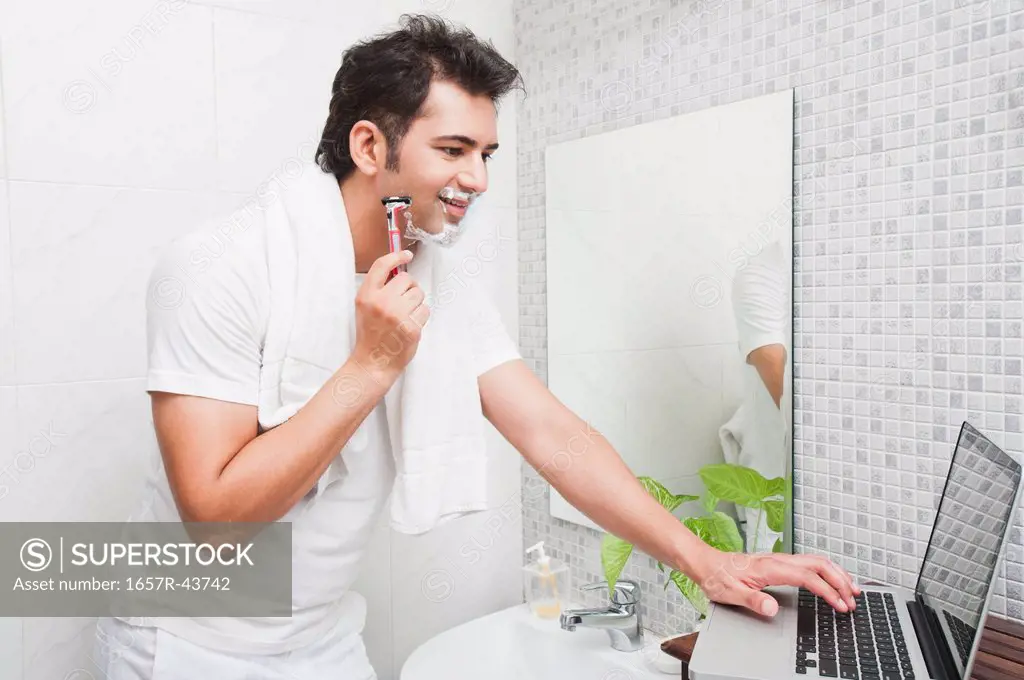 Man using a laptop while shaving his face