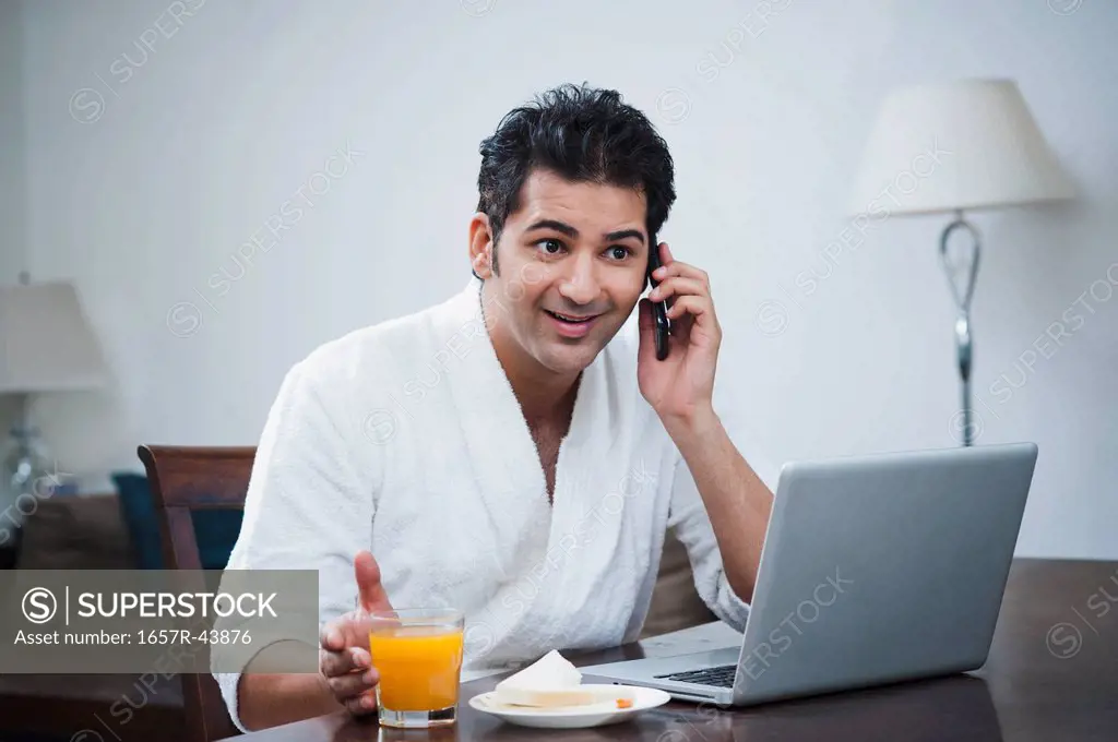 Businessman sitting in front of a laptop and talking on a mobile phone at breakfast table