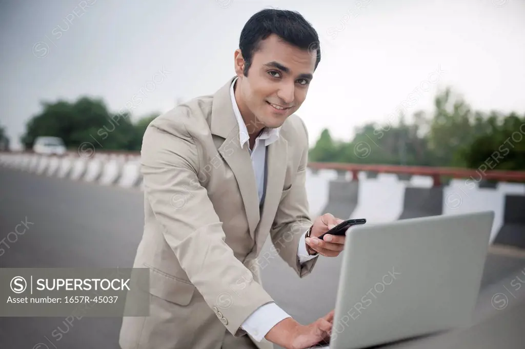Businessman text messaging while using a laptop on car bonnet at a flyover